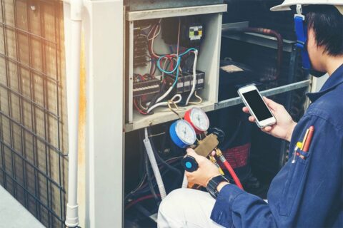 A man working on an air conditioning unit with a cell phone.