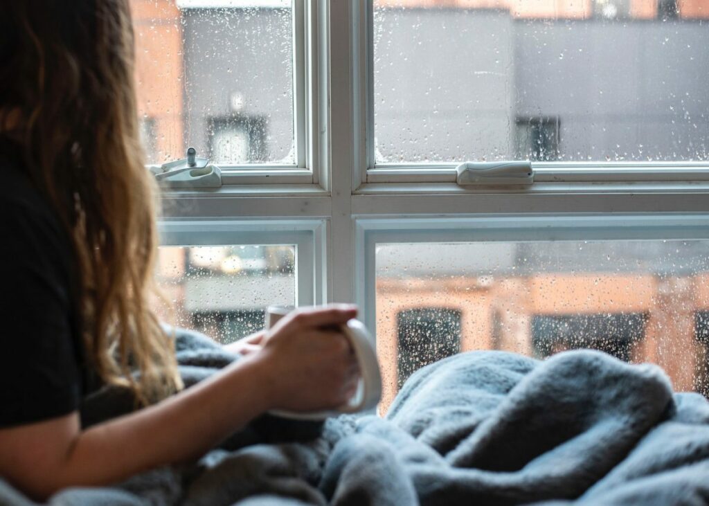 A woman is sitting on a bed with a cup of coffee in front of a window.