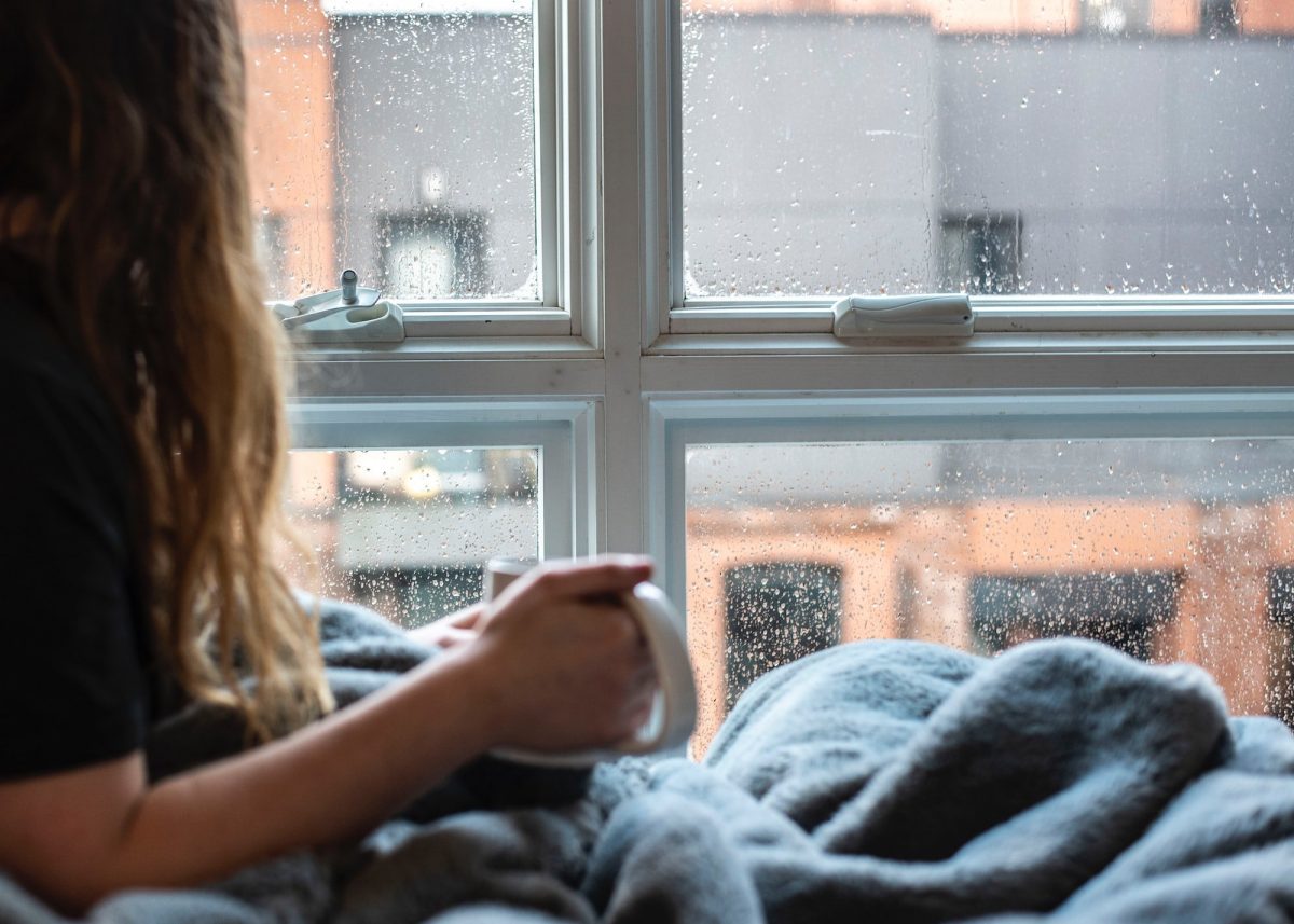 A woman is sitting on a bed with a cup of coffee in front of a window.