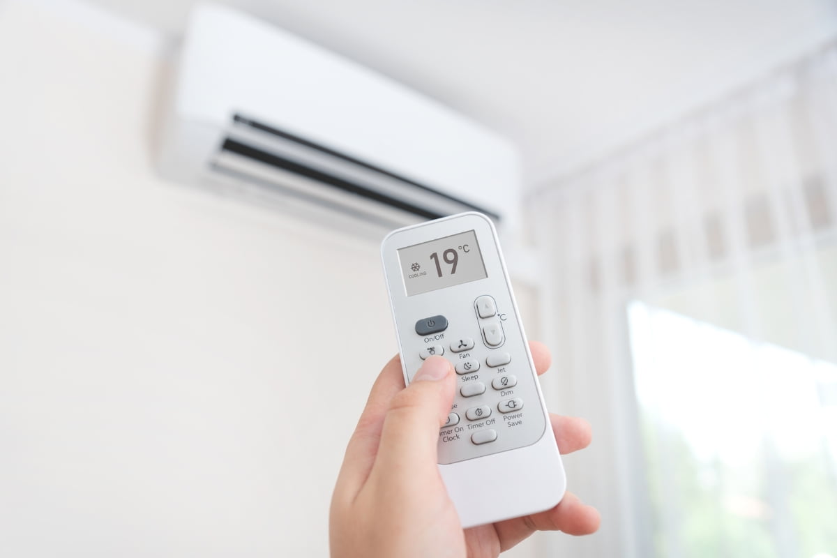 A person holding a wireless thermostat in front of an air conditioner.