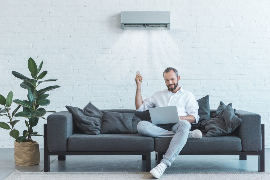 A man sitting on a couch with a laptop in front of a wireless thermostat.