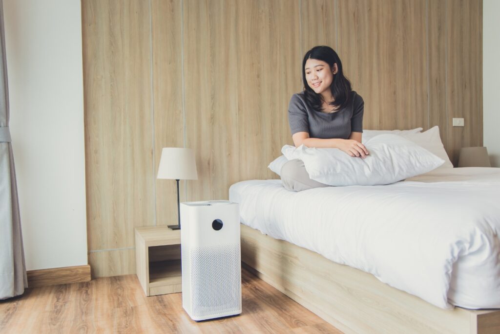 A woman sitting on a bed, taking care of her indoor air quality with an air purifier.