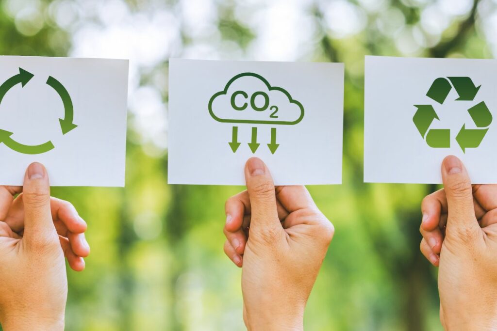 Three hands holding up cards with symbols for recycling, carbon emissions, and climate change against a blurred natural background.