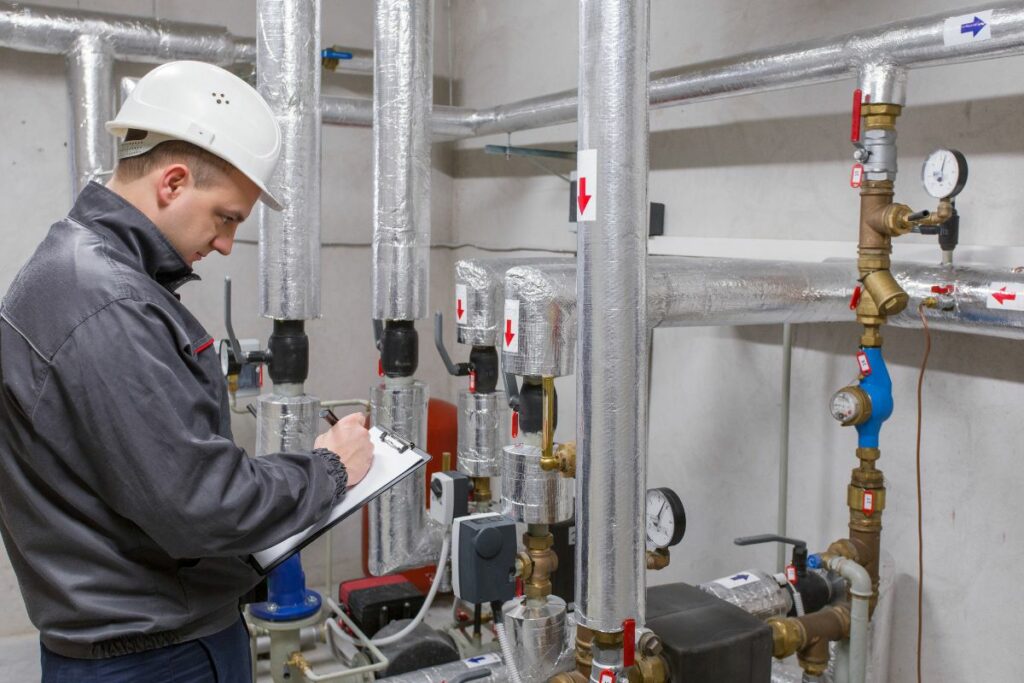 A technician in a hard hat taking notes on a clipboard while doing HVAC system analysis of pipes and valves in a boiler room.