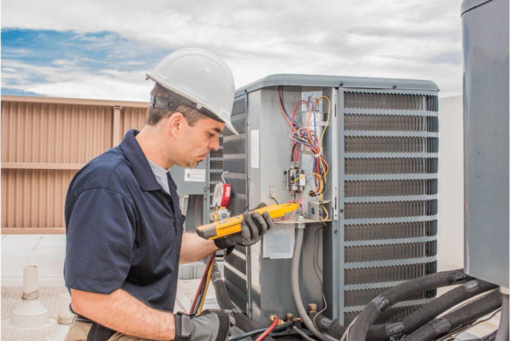 A technician in a hard hat uses a multimeter to inspect a new AC unit under a cloudy sky.