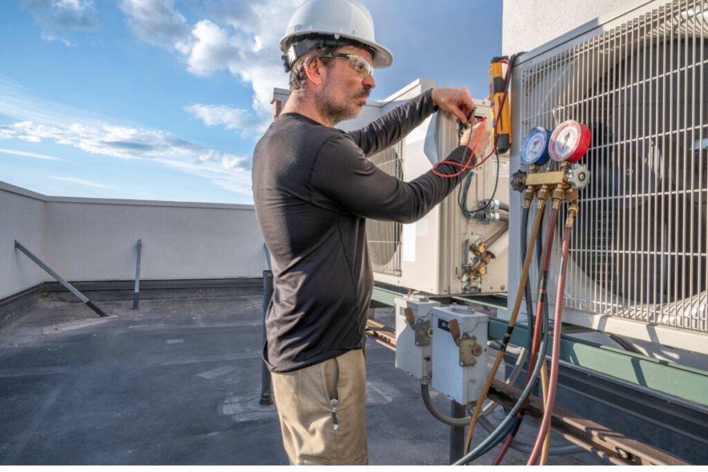 Technician with a helmet adjusting the gauges on a new AC unit on a rooftop, clear blue sky in the background.