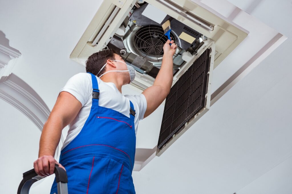 A technician in overalls and goggles installs a new AC unit on a ceiling, using a tool.