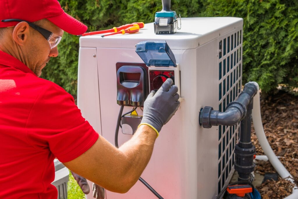 A technician in a red shirt and cap installs a new outdoor air conditioning unit, using tools placed nearby.