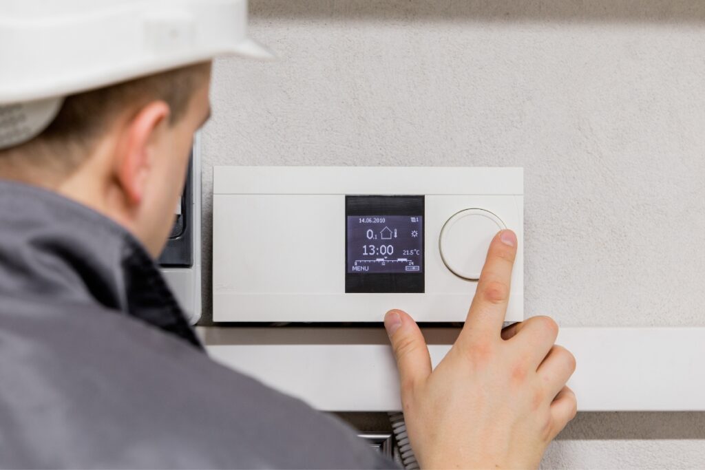 A person wearing a white hard hat adjusts a white digital thermostat on a wall, showcasing emerging HVAC technologies. The display shows temperature settings and time.