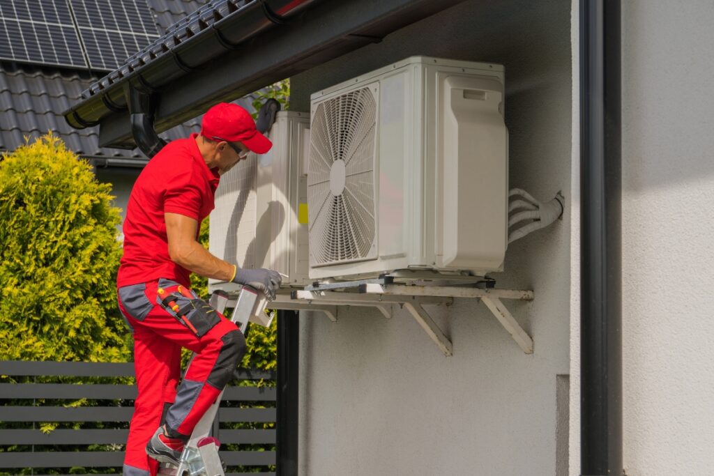 A technician in a red uniform and cap performs maintenance on an outdoor air conditioning unit mounted on a residential wall, utilizing emerging HVAC technologies.