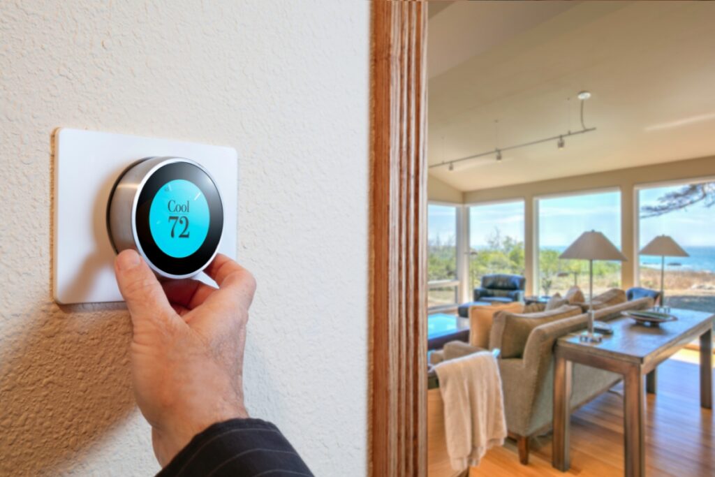 A person adjusts a digital thermostat set to 72 degrees Fahrenheit in a modern living room with large windows and a scenic view, embracing emerging HVAC technologies.