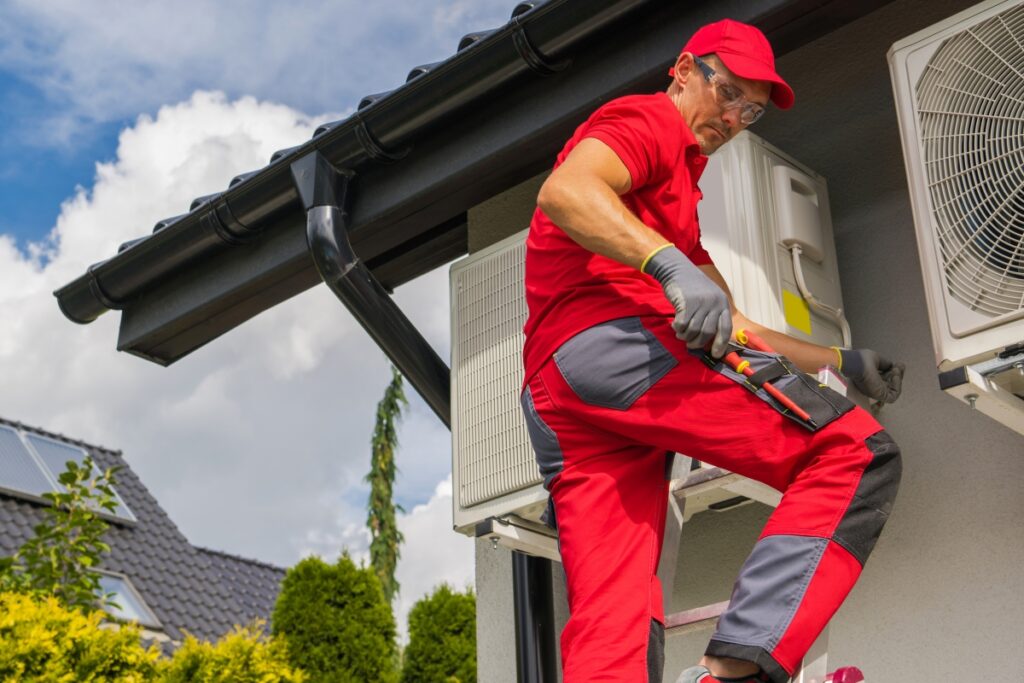 Technician in red uniform and cap, working on an outdoor air conditioning unit, holding a screwdriver, with a house roof and a cloudy sky in the background, demonstrating expertise in emerging HVAC technologies.