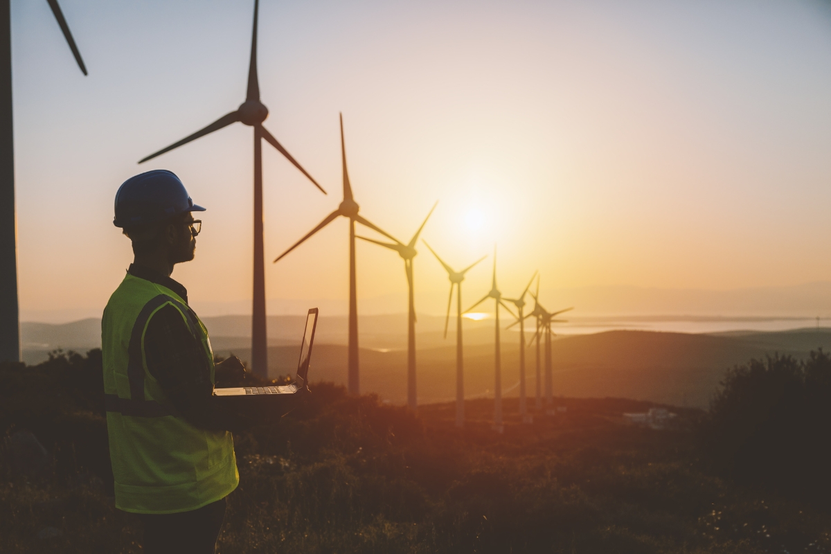 A person in a helmet and safety vest works on a laptop near wind turbines at sunset, monitoring renewable energy systems.