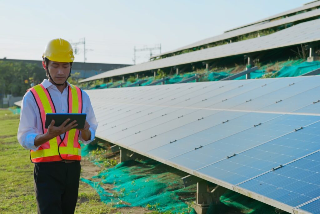 A person in a yellow construction helmet and reflective vest stands next to a large array of solar panels, examining a tablet, ensuring the efficiency of renewable energy systems.