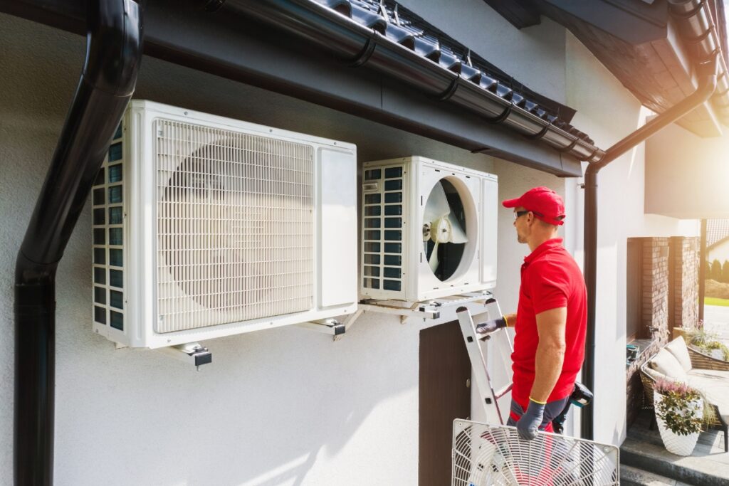 A technician in a red uniform and cap stands on a ladder next to two air conditioning units mounted on an exterior wall, performing maintenance or installation work on the HVAC system.