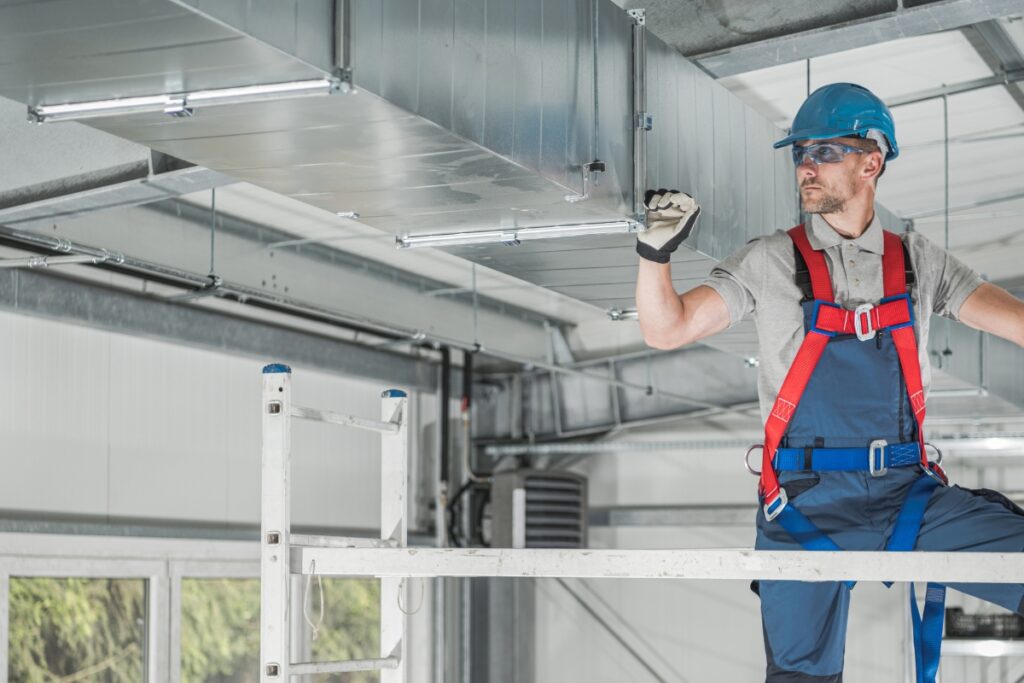 A construction worker wearing protective gear and a safety harness inspects ductwork on a raised platform inside an industrial building, ensuring the HVAC system integrates smoothly with renewable energy systems.