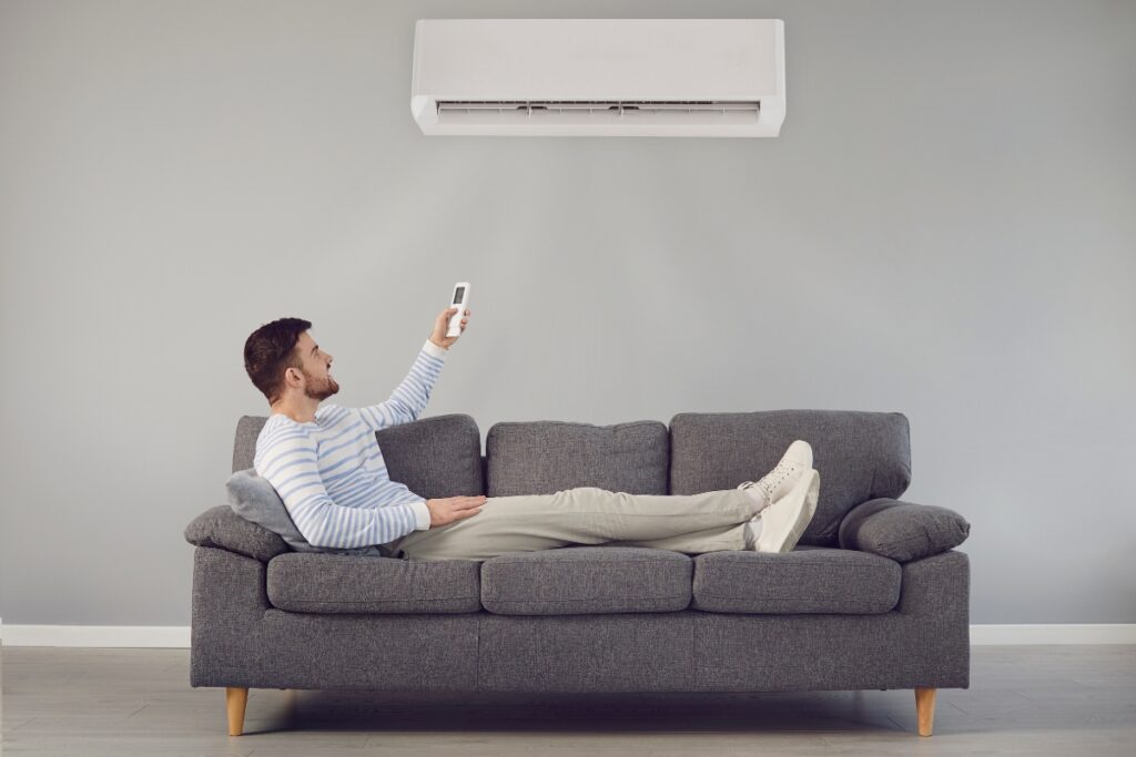 A man reclining on a gray couch uses a remote control to adjust the wall-mounted air conditioner above him, enjoying the health benefits of air conditioning in his comfortable living space.