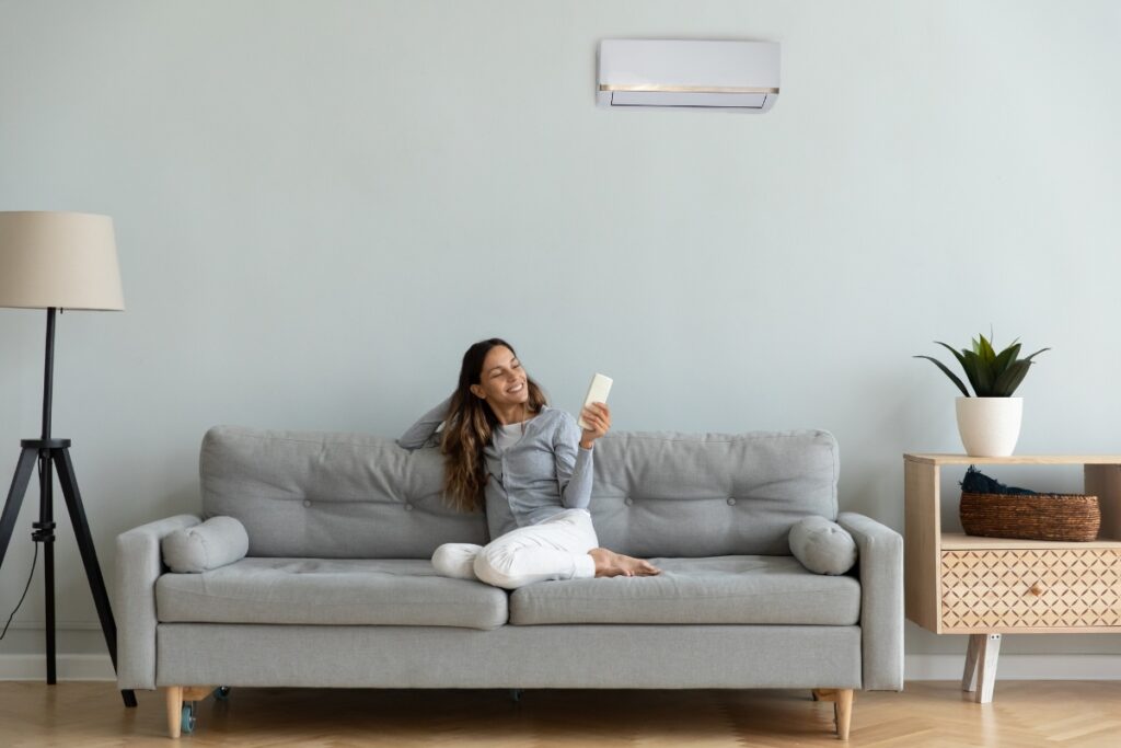 A woman sits on a gray couch with remote control, smiling, below a wall-mounted air conditioner in a minimalist living room, enjoying the health benefits of air conditioning.