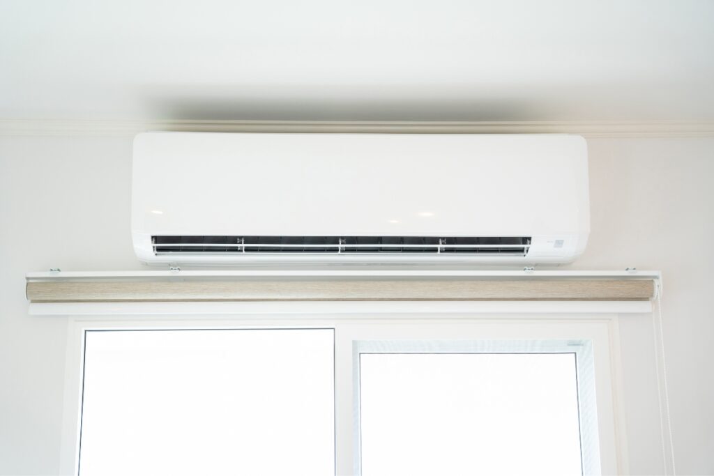 A white wall-mounted air conditioner installed above a window with a light-colored blind offers not only comfort but also the health benefits of air conditioning, helping to filter out allergens and maintain optimal indoor air quality.