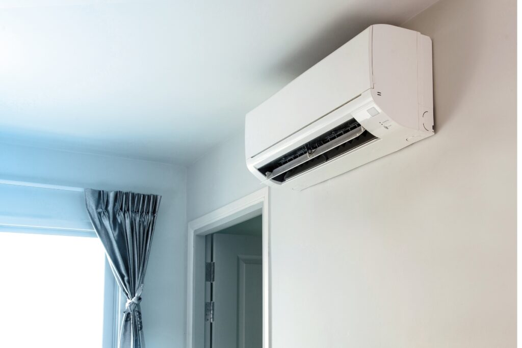 A wall-mounted air conditioning unit is installed near a door and a window with a blue curtain in a room with white walls, offering the added health benefits of air conditioning by maintaining an optimal indoor climate.