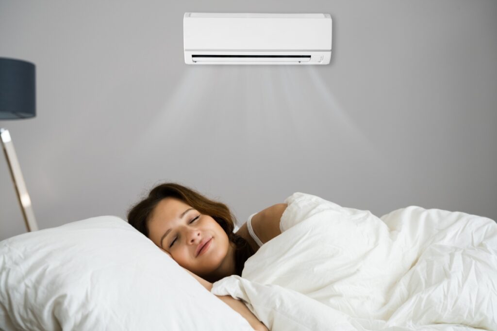 A woman sleeps peacefully in bed under a white duvet, enjoying the health benefits of air conditioning as a wall-mounted unit blows cool air above her.