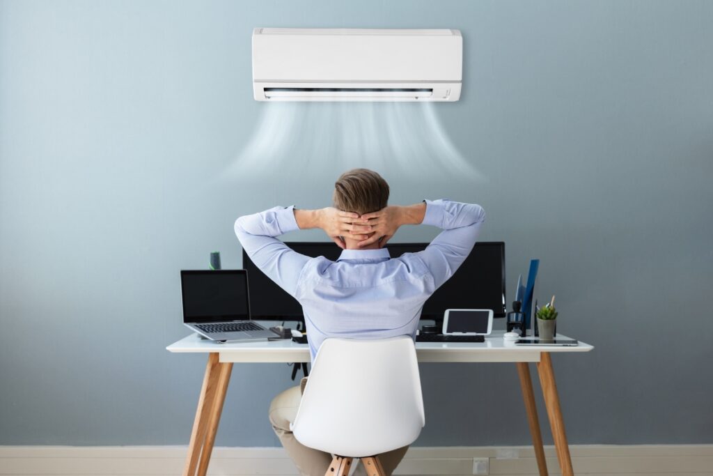         Person sitting at a desk with multiple screens and a laptop, enjoying the health benefits of air conditioning from a unit mounted on the wall above.