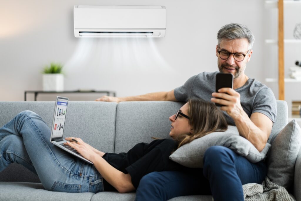 A man and woman are on a grey sofa; the man is using a smartphone while the woman, with her head resting on a pillow, is using a laptop. An air conditioner, providing health benefits of air conditioning, is mounted on the wall behind them.