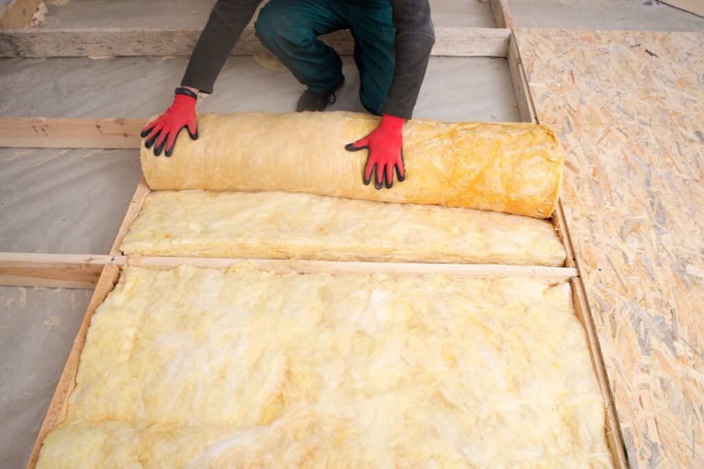 A person wearing red gloves is installing yellow fiberglass insulation in a floor frame, ensuring meticulous hvac planning for optimal energy efficiency.