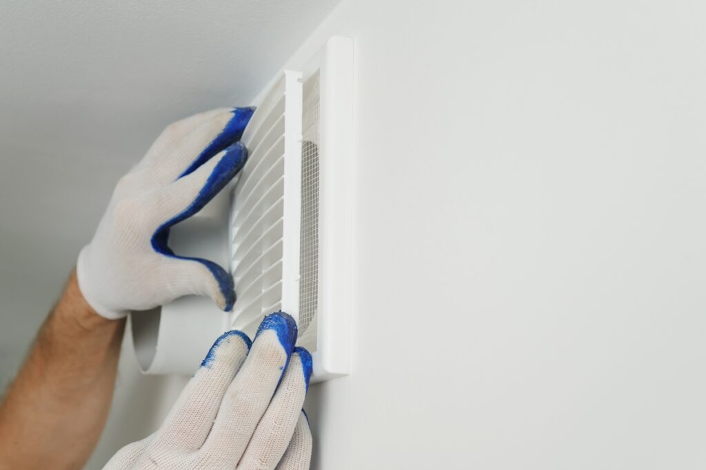 A person wearing white gloves with blue fingertips is installing or adjusting a white air vent cover on a white wall, demonstrating meticulous attention to detail in HVAC planning.