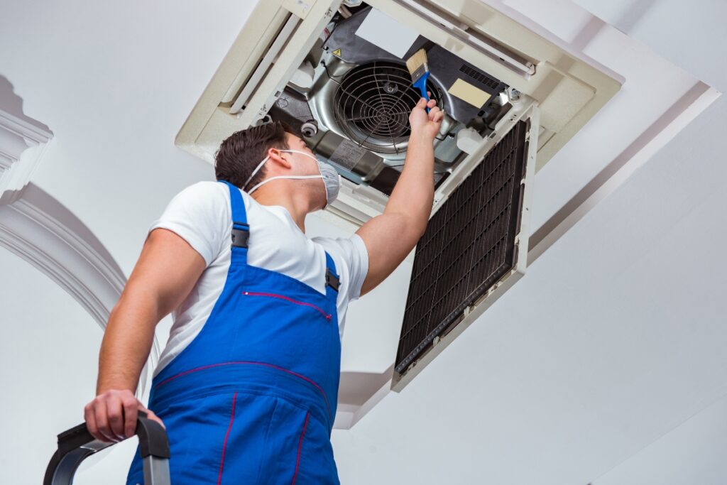 A technician in blue coveralls and a mask, meticulously adhering to hvac planning, is cleaning the inside of a ceiling air conditioner unit with a brush while standing on a ladder.