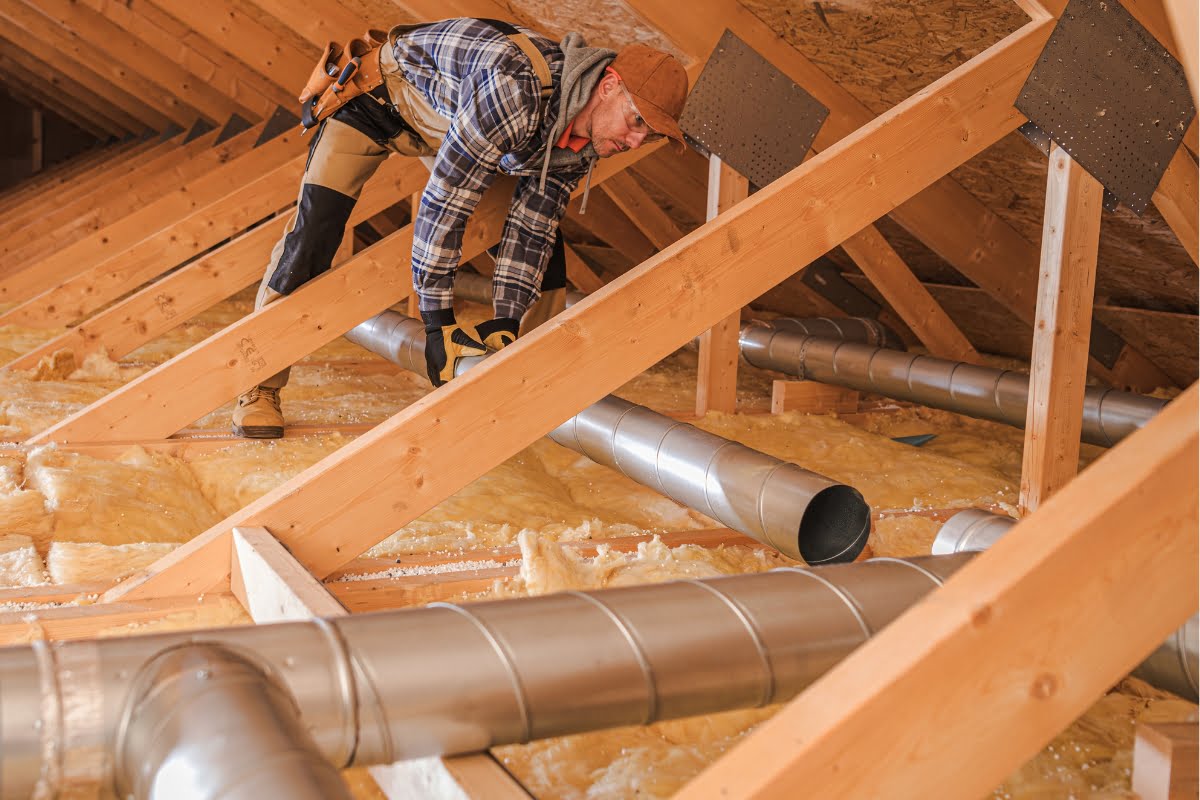 A worker in a flannel shirt and cap installs HVAC ducts in an attic, surrounded by wooden beams and insulation material.