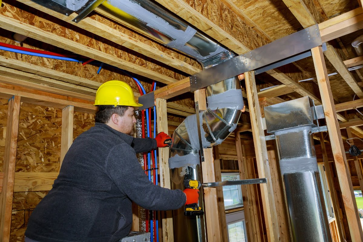 A construction worker wearing a yellow hard hat and gloves is using a power drill to install ductwork and HVAC insulation in a wooden-framed building.