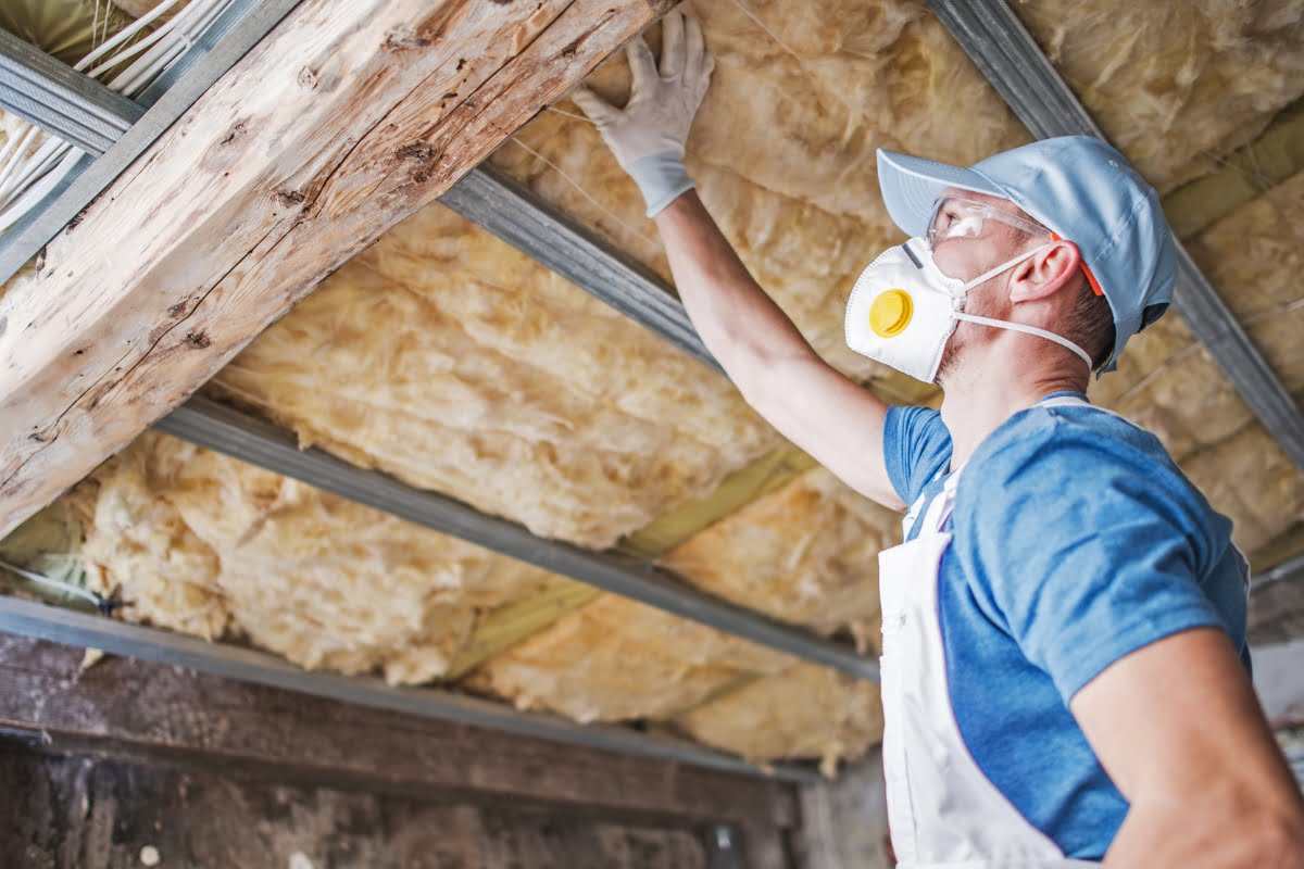 A worker in a blue shirt, white overalls, and a cap installs hvac insulation in the ceiling. The worker is wearing a protective mask and gloves.