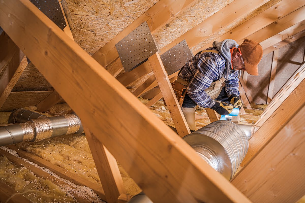 A person in work clothes and a cap is inspecting and working on the HVAC ductwork in an attic with wooden beams, ensuring the insulation is properly installed.