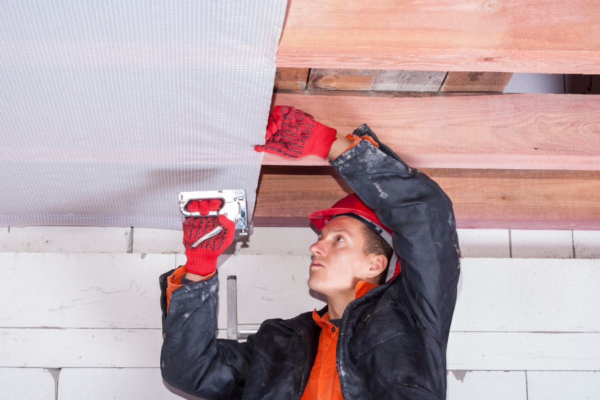 A construction worker wearing protective gear staples HVAC insulation material to the ceiling framework of a building.