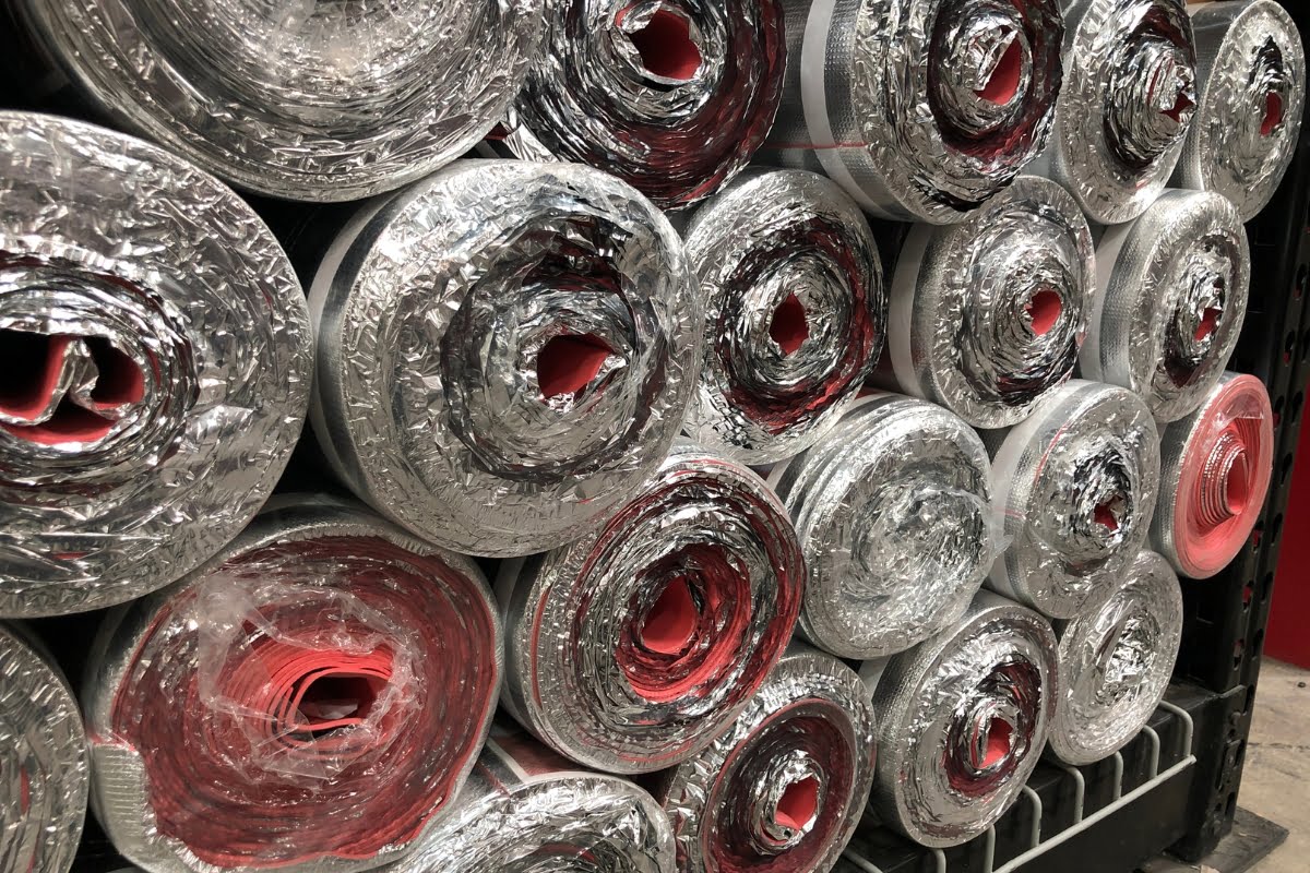 Close-up of stacked rolls of HVAC insulation material with a silver outer layer and red-colored inner layer, stored on metal racks.