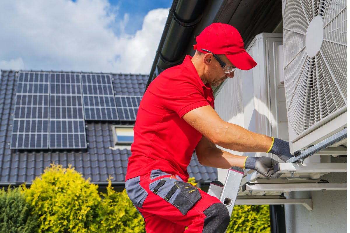 A technician in a red uniform works on a wall-mounted air conditioning unit outside a building with solar panels on the roof, ensuring proper HVAC insulation.