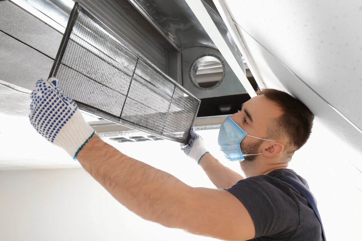 A person wearing gloves and a mask is installing or removing a filter from an overhead ventilation system, ensuring proper HVAC insulation.