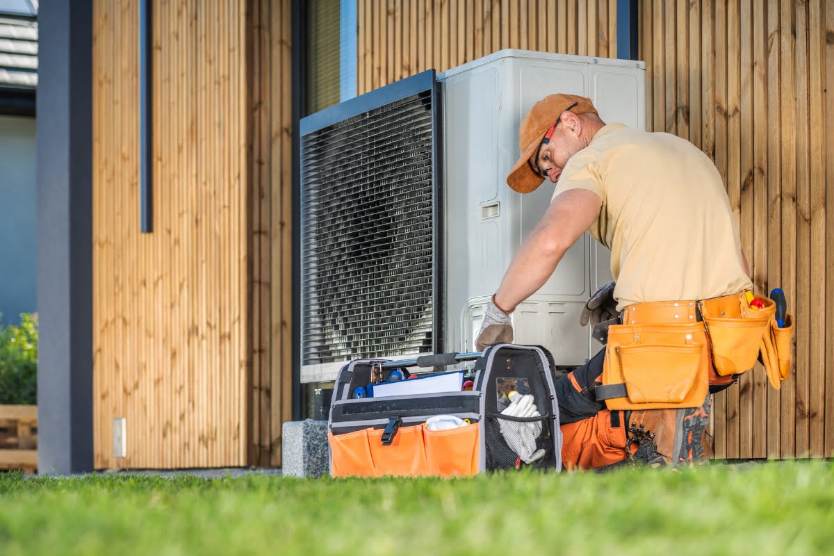 A technician wearing an orange cap and tool belt works on an outdoor air conditioning unit beside a wooden building. In the well-maintained hvac landscaping, a tool bag is open on the grass near him.