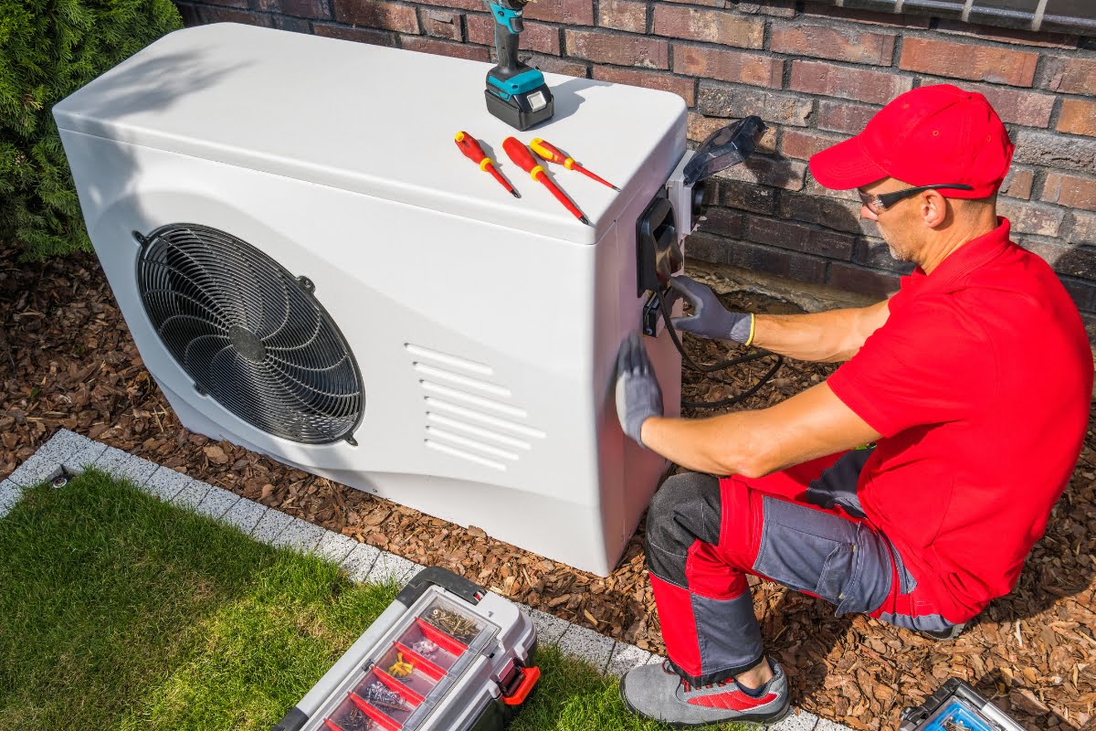 A technician in a red uniform installs or repairs an outdoor air conditioning unit, surrounded by tools on top and a toolbox nearby, while mindful of landscaping for energy efficiency.