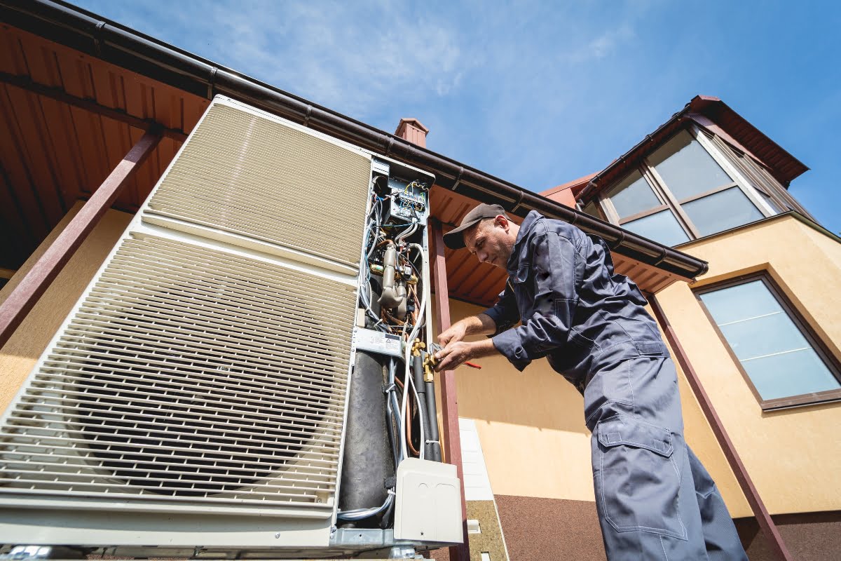 A technician in a gray uniform is repairing an outdoor air conditioning unit attached to a building under a clear sky, amid carefully planned HVAC landscaping for energy efficiency.