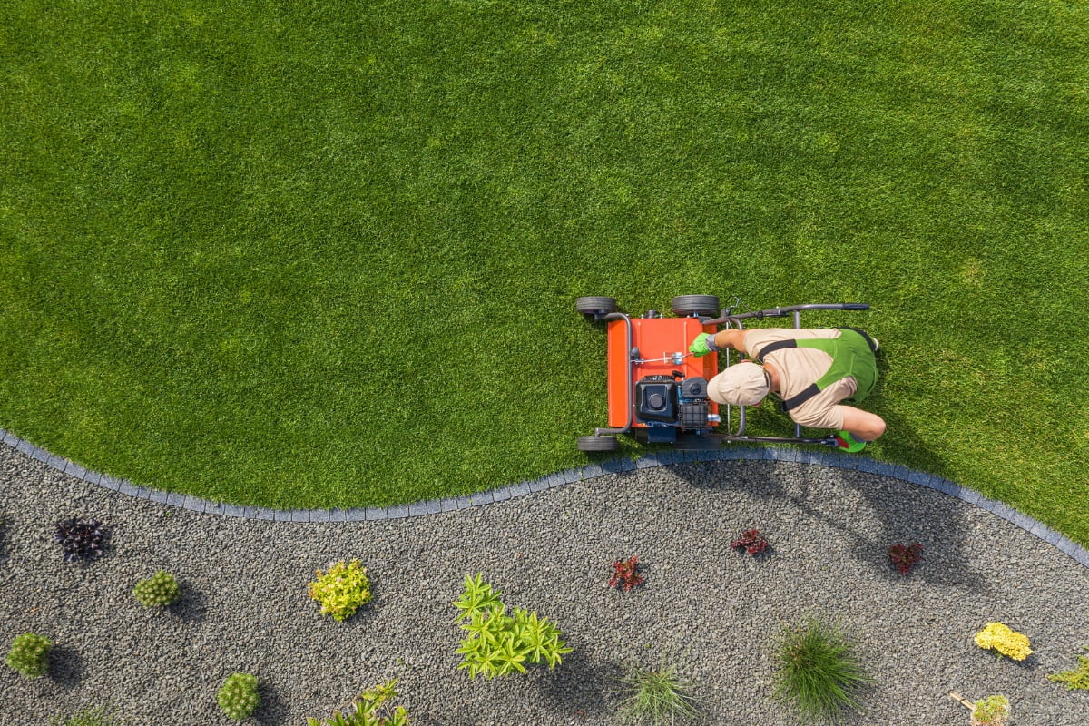 A person in green overalls is pushing a red lawnmower, skillfully cutting the grass along a curved edge that separates a gravel area with small plants. This scene highlights how landscaping for energy efficiency can create visually appealing and functional outdoor spaces.