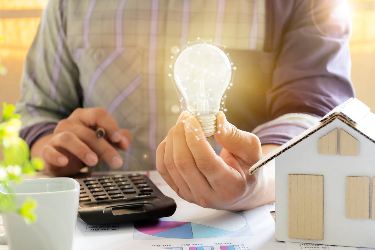 A person holding a glowing light bulb sits at a desk with a calculator, a small model of a house, financial documents, and brochures on landscaping for energy efficiency.