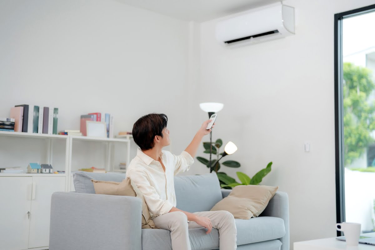 A person is sitting on a couch using a remote control to operate a wall-mounted air conditioner in a bright, modern living room, knowing that hvac emergency services are just a call away.