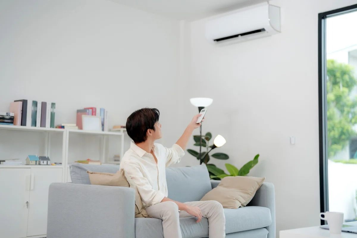 A person is sitting on a couch using a remote control to operate a wall-mounted air conditioner in a bright, modern living room, knowing that hvac emergency services are just a call away.