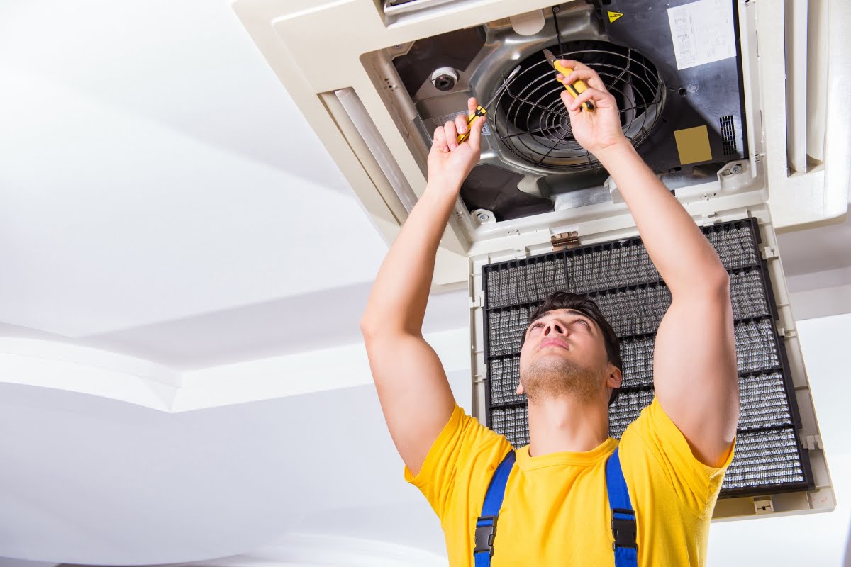 A technician in a yellow shirt and blue overalls is repairing a ceiling air conditioning unit, using tools to work on components inside the open unit as part of HVAC emergency services.