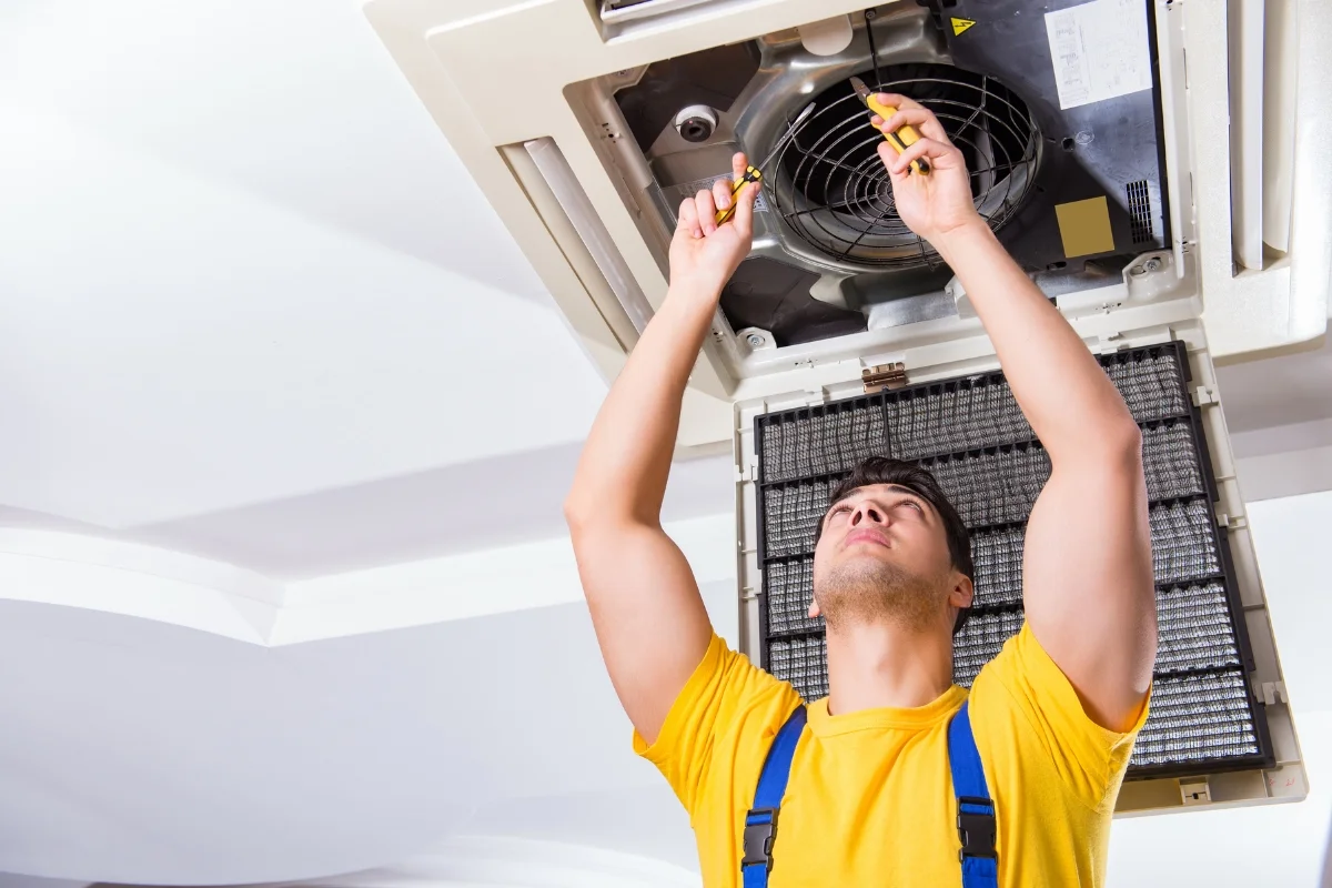 A technician in a yellow shirt and blue overalls is repairing a ceiling air conditioning unit, using tools to work on components inside the open unit as part of HVAC emergency services.