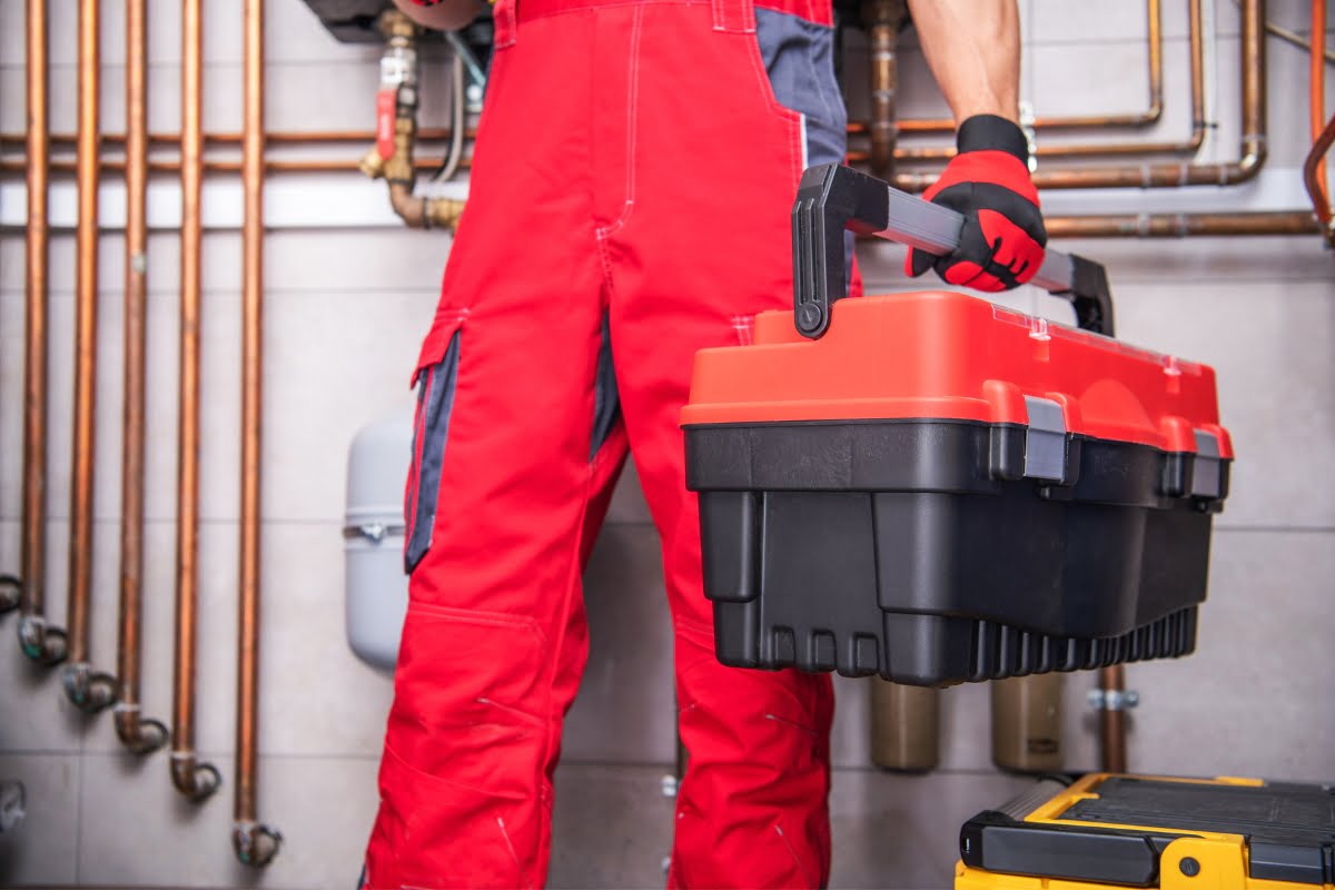 A worker in red overalls, holding a black and red toolbox, stands in front of pipes and valves in a mechanical or plumbing setting, ready to provide HVAC emergency services.