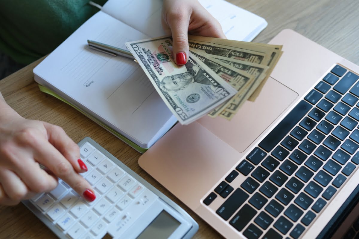 A person with red nail polish calculates expenses with a calculator while holding U.S. dollar bills, near a laptop and notebook on a wooden desk, perhaps tallying up costs for recent HVAC emergency services.