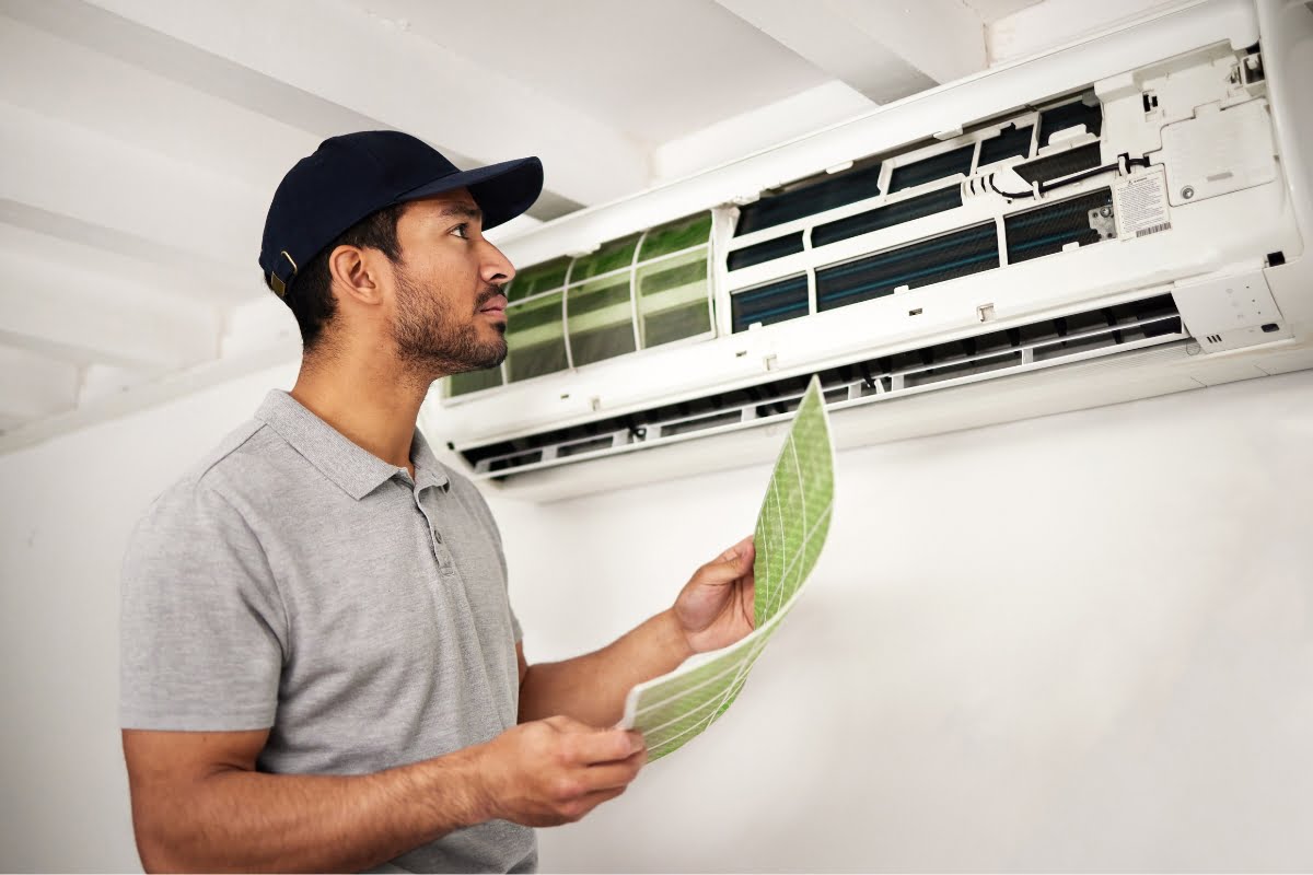 A man in a gray shirt and cap inspects and replaces the filters of a wall-mounted air conditioner, showcasing his expertise in HVAC emergency services.